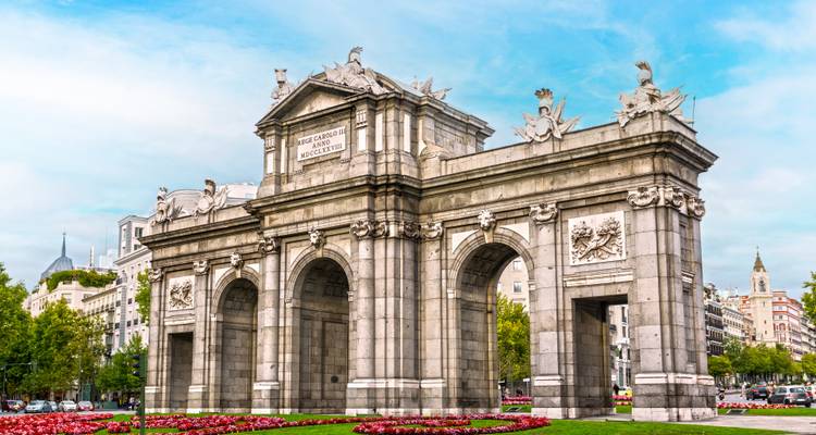 Stone triumphal arch Puerta de Alcalá surrounded by flowerbeds and city buildings under blue sky.
