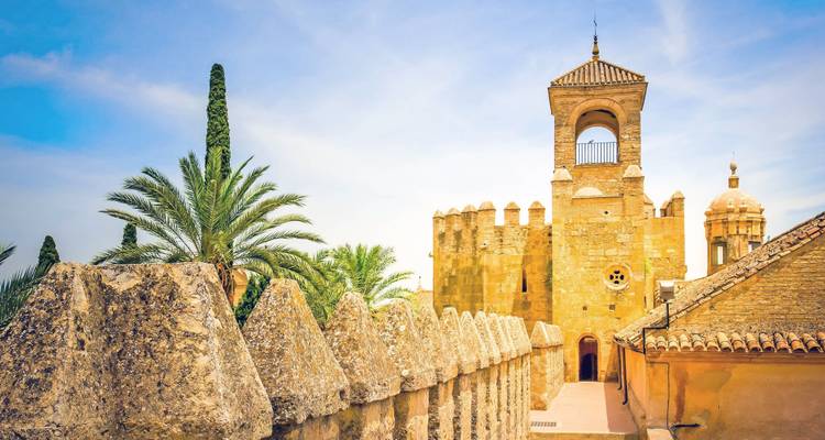 Golden stone tower and crenellations of Cordoba’s Alcázar with palm trees and battlements in warm light.
