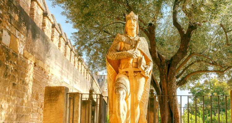 Stone statue of a medieval knight in Cordoba’s Alcázar gardens under an olive tree.