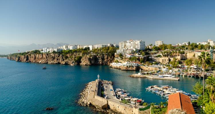 Vue depuis une falaise surplombant une baie turquoise, un vieux port, un phare et les bâtiments blancs d'une ville méditerranéenne.