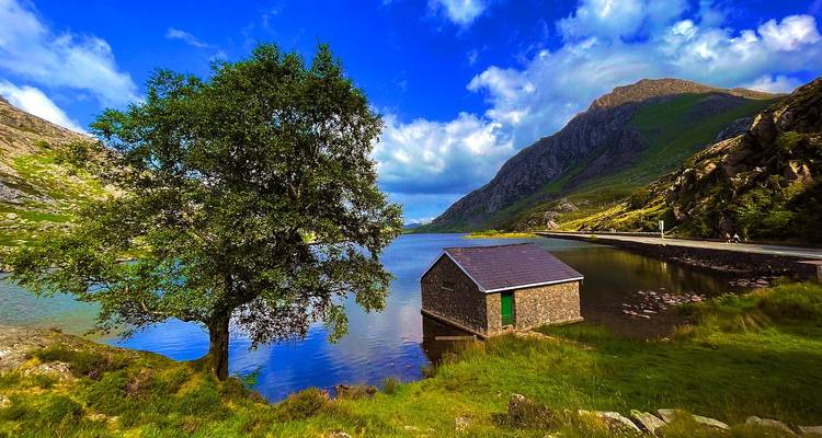 Scène de lac vivante avec un hangar à bateaux en pierre, un arbre et des montagnes sous un ciel bleu éclatant