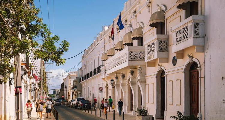 Pedestrians strolling along a charming colonial street with white façades, balconies and flags.