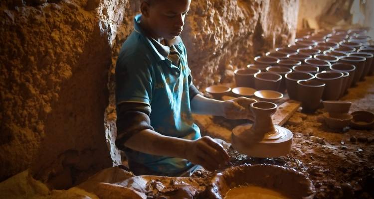 Un potier façonnant l'argile sur un tour traditionnel à l'intérieur d'un atelier faiblement éclairé bordé de pots frais