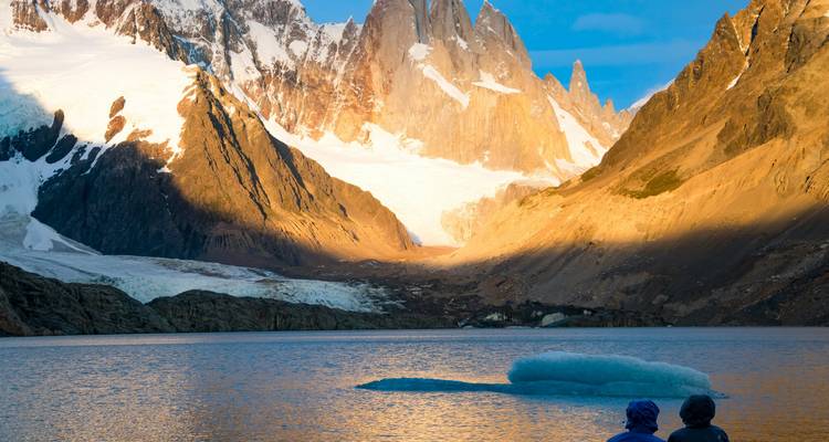 Goldenes Licht badet die gezackten Gipfel des Mount Fitz Roy und einen Gletscher, der in einen türkisfarbenen See fließt, wo zwei Wanderer die Aussicht bewundern.
