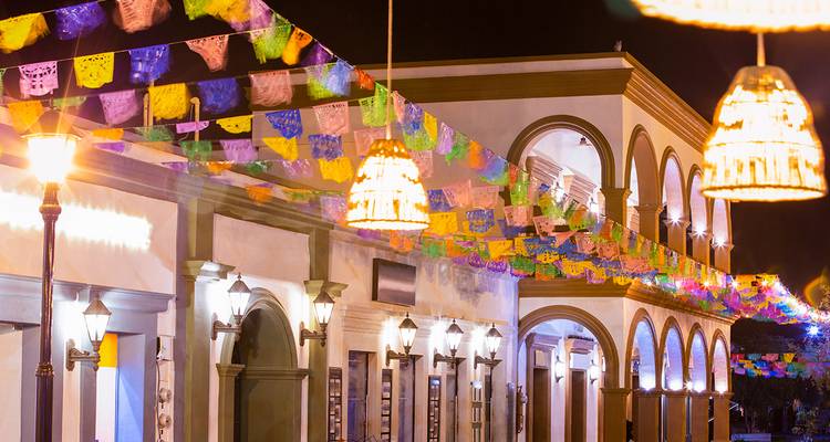 Une rue coloniale festive la nuit décorée de drapeaux colorés en papel picado et d'une lumière chaude de lampadaires.