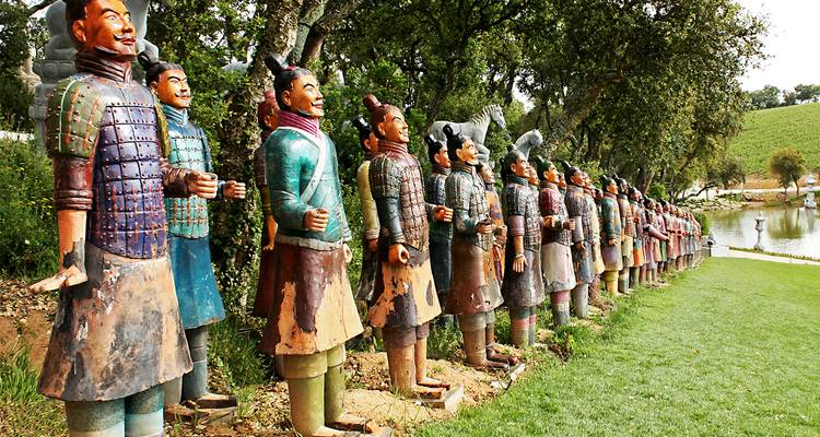 A long line of brightly painted Terracotta Warrior statues stands beside a grassy pond under leafy trees.