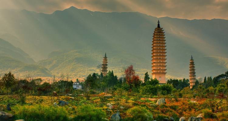 Three ancient pagodas rise from lush gardens with dramatic mountains behind, lit by soft golden light streaming through clouds.