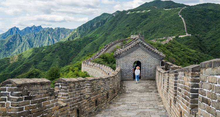 Ein Besucher geht entlang einer steinernen Brüstung der Chinesischen Mauer, die sich über üppige Bergkämme erstreckt