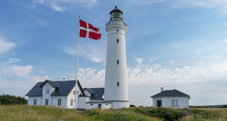 White lighthouse with Danish flag standing in grassy dunes under a pale blue sky.