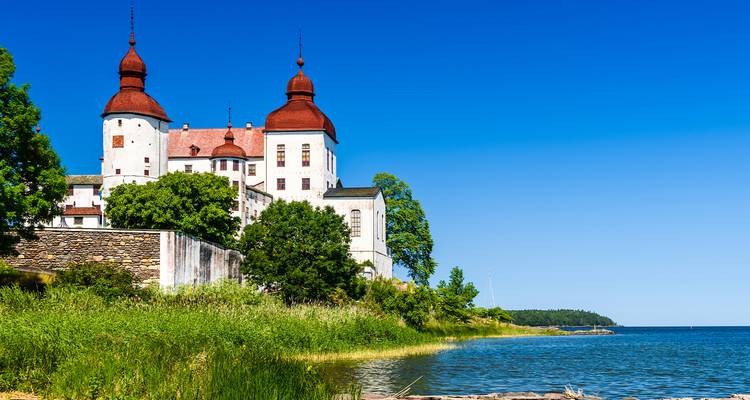 Historic lakeside castle with red towers rises above lush greenery against a deep blue sky.