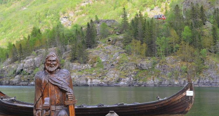Wooden Viking longship replica and carved warrior statue beside a still fjord backed by forested cliffs.