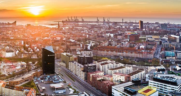 Urban panorama at sunrise with golden light illuminating a coastal Danish city and busy port cranes.