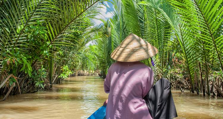Un voyageur portant un chapeau conique navigue en bateau en bois à travers les luxuriants canaux de palmiers du Delta du Mékong.