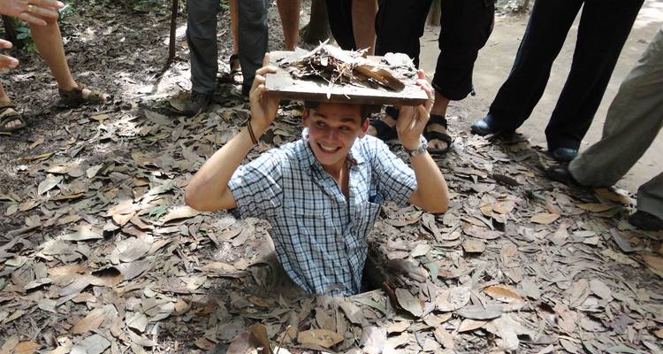 Un visiteur émerge en souriant d'une trappe étroite des tunnels historiques de Cu Chi, entouré d'un sol couvert de feuilles.