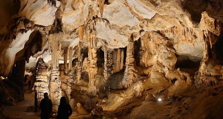 Weitläufige Kalksteinhöhlenkammer in Bernsteintönen beleuchtet mit Stalaktitensäulen und erkundenden Besuchern