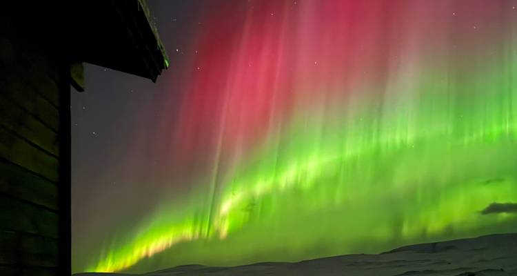 Vivid pink and green aurora sweeps over snowy hills beside the silhouette of a wooden cabin.
