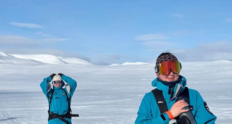 Two adventurers in teal snowsuits stand on a vast, windswept snow plateau under a blue Arctic sky.