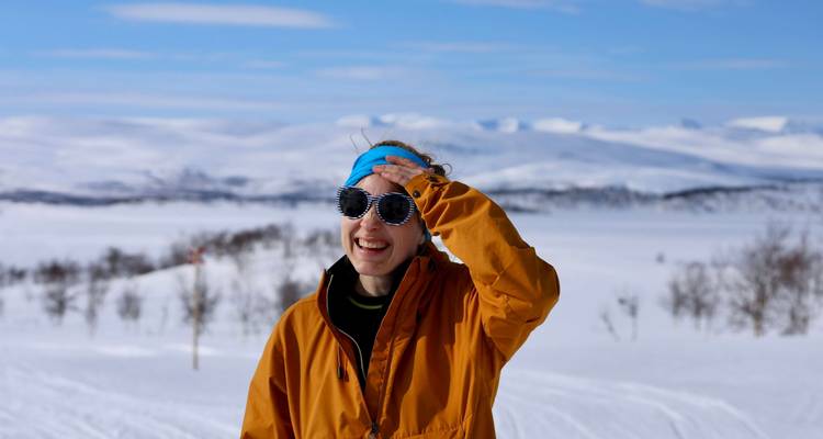 A smiling skier in a mustard jacket enjoys sunny alpine views over endless snowy hills.