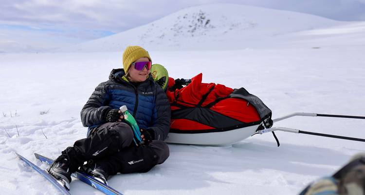 A skier rests on snow beside a red-packed pulk sled amid white Arctic wilderness.