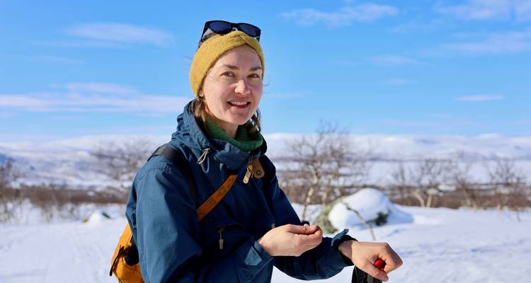 Young woman in winter trekking gear smiling while resting on snowy landscape with blue sky and distant hills.