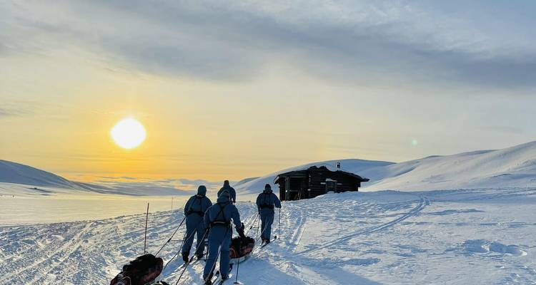 Group of skiers pulling sleds toward a remote cabin under a low golden sun on snowy plateau.