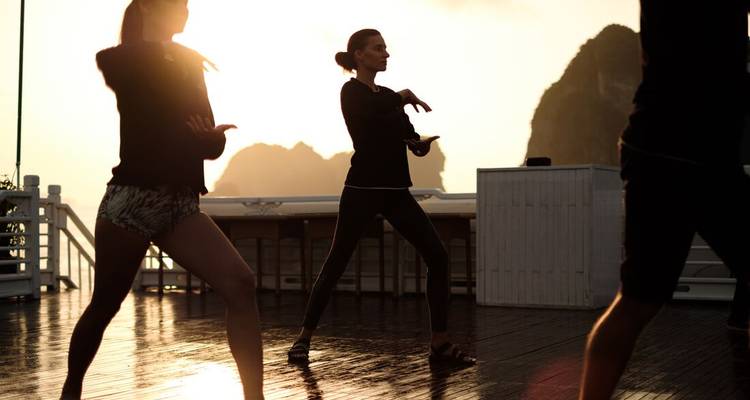 Los viajeros practican tai chi al amanecer en una cubierta de madera de un crucero por la Bahía de Halong con siluetas de formaciones rocosas de fondo.