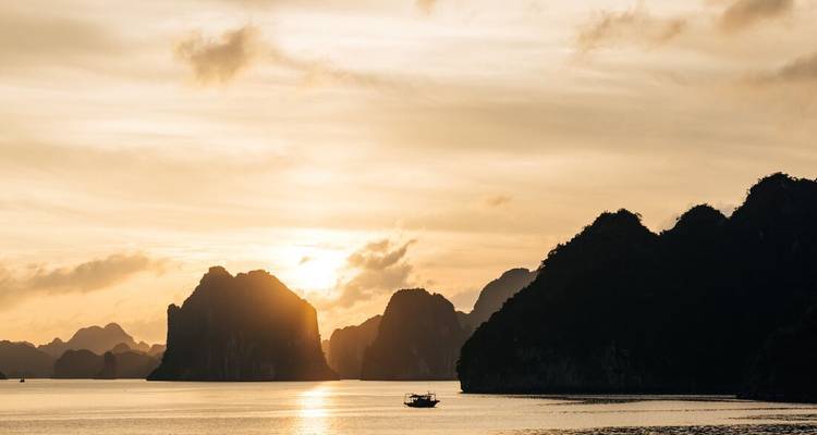 Atardecer dorado sobre la Bahía de Halong con un bote solitario recortado contra el agua tranquila e islas de piedra caliza dramáticas.