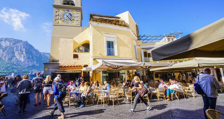 La plaza principal soleada de Capri con torre del reloj, terrazas de cafés llenas de vida y multitudes de visitantes.