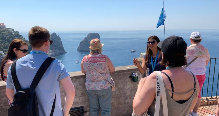 Guía hablando a turistas en una terraza con vistas a los Faraglioni de Capri y al mar azul profundo.
