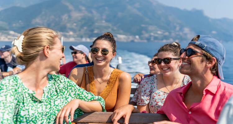 Grupo de jóvenes viajeros riendo en un ferry rápido con la costa montañosa de fondo.