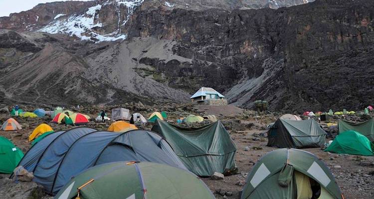 Groep gekleurde tenten opgezet op rotsachtige grond onder de steile rotswand van de Kilimanjaro.