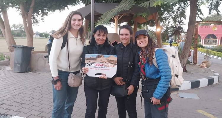 Quatre jeunes femmes portant des sacs à dos posent à l'extérieur en tenant une pancarte pour leur circuit touristique en Égypte.