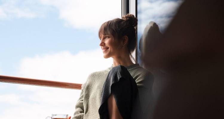 Une jeune femme sourit en savourant une boisson chaude sur le balcon d'une croisière fluviale avec un ciel en arrière-plan.