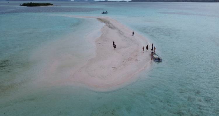 Un petit banc de sable émerge dans une eau turquoise peu profonde où plusieurs voyageurs et un bateau sont amarrés.