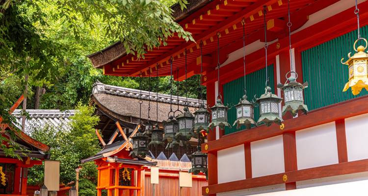 Close-up of hanging lanterns and vermilion beams of traditional Shinto shrine among greenery.