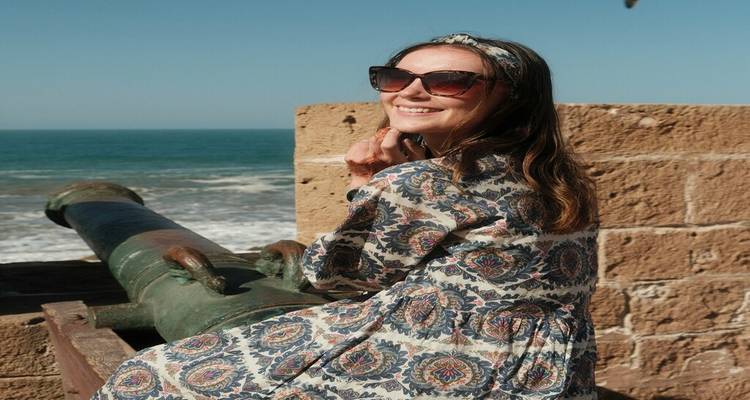 Femme souriante vêtue d'une robe à motifs assise sur un ancien canon au sommet de remparts en bord de mer avec des vagues qui s'écrasent ci-dessous.