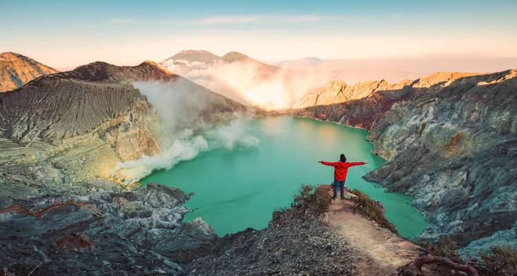 Un randonneur en veste rouge se tient les bras écartés au-dessus d'un lac de cratère turquoise entouré de crêtes volcaniques escarpées à l'aube.