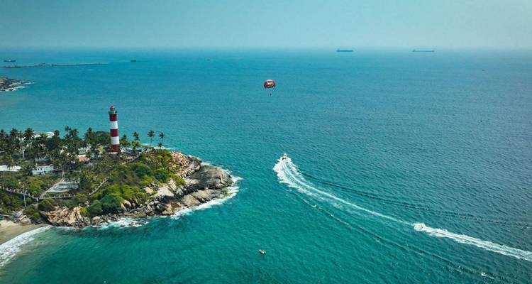 Luchtfoto van een rood-wit gestreepde vuurtoren op een rotsachtige kaap met een parasailer en motorbootjes die witte golven achterlaten in de turquoise zee.
