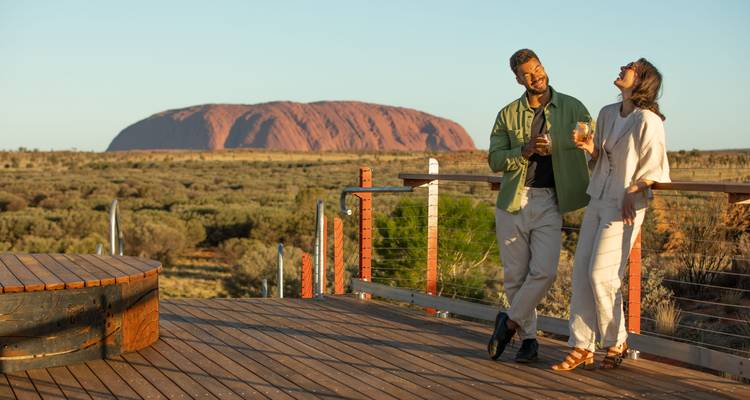 Couple dégustant des boissons sur une terrasse en bois avec Uluru brillant de lumière dorée en arrière-plan.