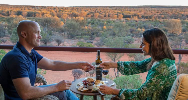 Un couple partage des amuse-bouches et du vin sur un balcon surplombant une brousse désertique rouge au crépuscule.