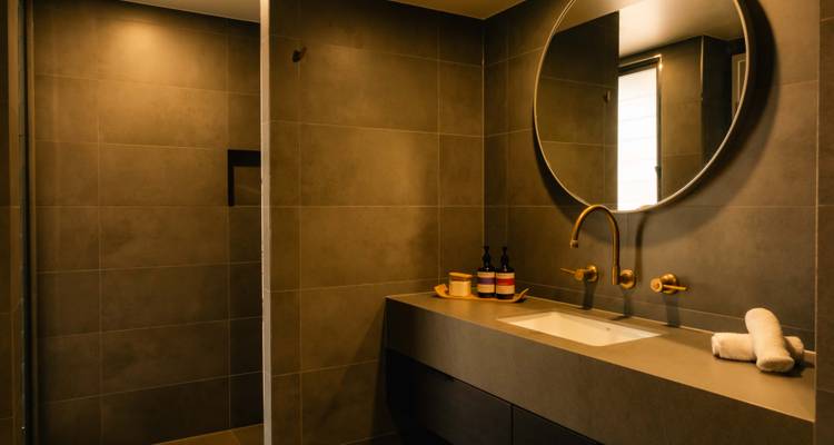 Modernes Badezimmer mit großem rundem Spiegel, grauen Fliesen und Messingarmaturen.