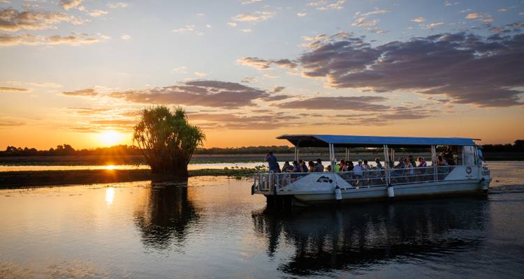 Ausflugsboot fährt bei Sonnenuntergang über ein ruhiges Billabong, wobei sich der goldene Himmel im Wasser spiegelt.
