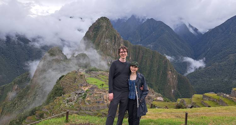 Un couple se tient devant les ruines brumeuses du Machu Picchu et les montagnes vertes escarpées sous un ciel nuageux.