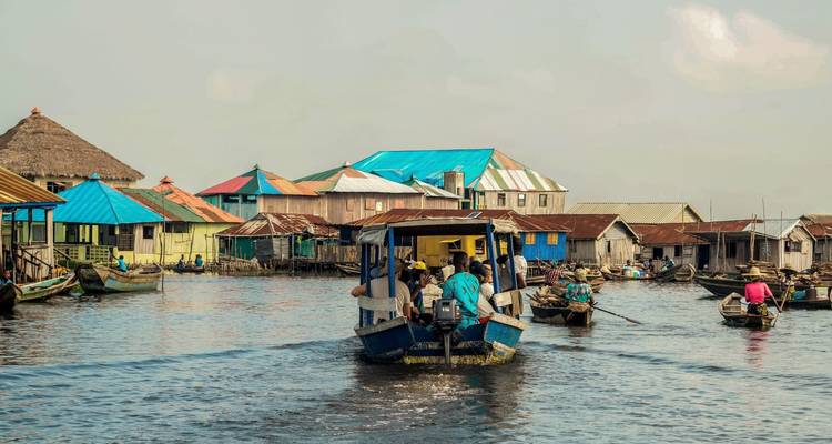 Des bateaux naviguant à travers un village sur pilotis avec des maisons aux toits en tôle colorée sur un large lagon.