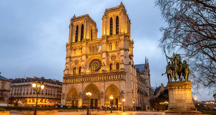 Cathédrale Notre-Dame illuminée au crépuscule avec des lampadaires lumineux et une statue à proximité.