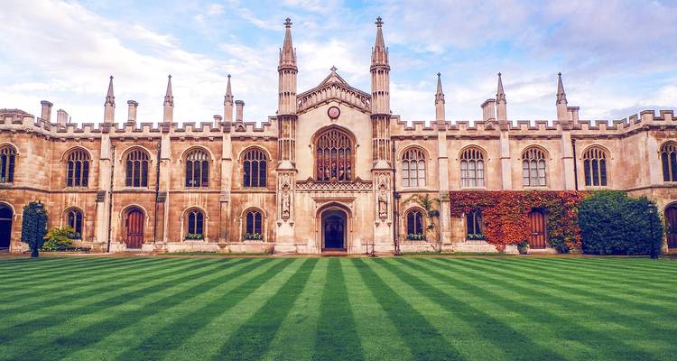 Elegant neo-gothic college building with spires faces a perfectly striped green lawn under a blue sky in Cambridge.