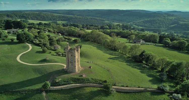 Drone view of a solitary stone tower atop rolling green hills and winding paths in the Cotswolds countryside.