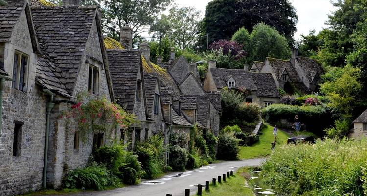 Quintessential Cotswold stone cottages with steep roofs line a narrow lane adorned with hanging flowers and greenery.