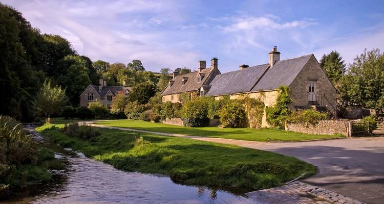 Stone cottages stand beside a gentle stream and lush lawns under a bright blue English sky.