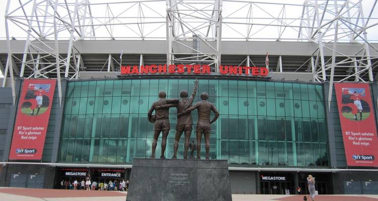 Statue de trois légendes du football face à l'entrée du stade Old Trafford avec une signalisation audacieuse en rouge « Manchester United ».